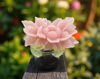 Peut inclure: Une sculpture détaillée d'une fleur rose, posée sur une base transparente et noire. La sculpture est exposée sur une surface en bois avec un arrière-plan flou vert et coloré.