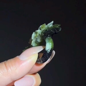 以下が含まれることがあります： 花柄のデザインが施された、彫刻された翡翠の指輪。指輪は、上部に明るい緑と白の花の彫刻が施された、濃い緑と黒のバンドが付いています。指輪は暗い背景に対して保持されています。