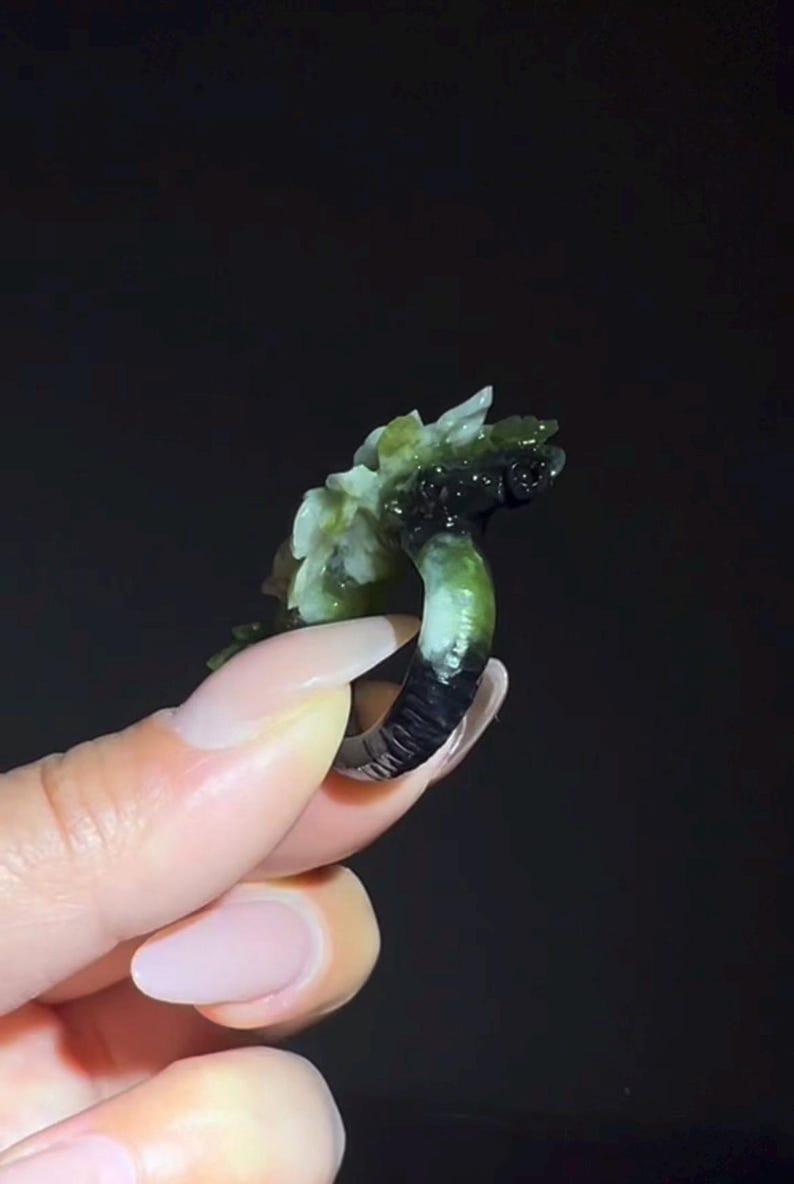 以下が含まれることがあります： 花柄のデザインが施された、彫刻された翡翠の指輪。指輪は、上部に明るい緑と白の花の彫刻が施された、濃い緑と黒のバンドが付いています。指輪は暗い背景に対して保持されています。