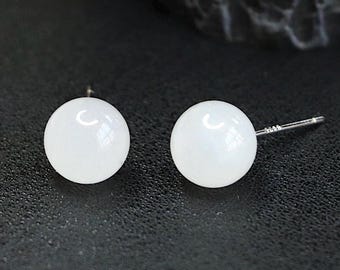 May include: A pair of white, round stud earrings. Each earring has a smooth, glossy surface and a silver post. The earrings are set against a dark background, highlighting their bright white color. The earrings are a classic jewelry item.
