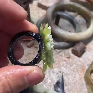 以下が含まれることがあります： 黒いリングと薄緑色の花の模様。リングは持ち上げられ、花と暗いバンドの複雑なディテールが示されています。他のリングが背景に見えます。