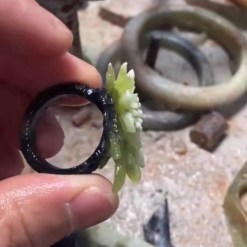 以下が含まれることがあります： 黒いリングと薄緑色の花の模様。リングは持ち上げられ、花と暗いバンドの複雑なディテールが示されています。他のリングが背景に見えます。