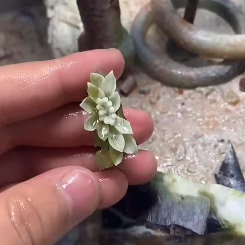 以下が含まれることがあります： 手で持たれた、彫刻された薄緑色の花の彫刻。彫刻は、繊細で半透明な外観の、詳細な花びらと葉を持っています。背景には、同じ素材の他の作品がある作業面が表示されています。