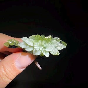 以下が含まれることがあります： 明るい緑と白のクリスタルフラワー彫刻。彫刻は、暗いベースで、複数の層の花びらと葉を持っています。クリスタルは黒い背景に対して手で保持されています。