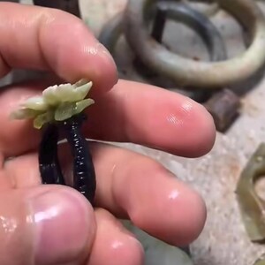 以下が含まれることがあります： 黒いリングに、彫刻された淡い緑色の花の模様。リングは手で持たれており、背景には他の翡翠のジュエリーが見えます。花には複数の花びらがあり、リングの焦点となっています。