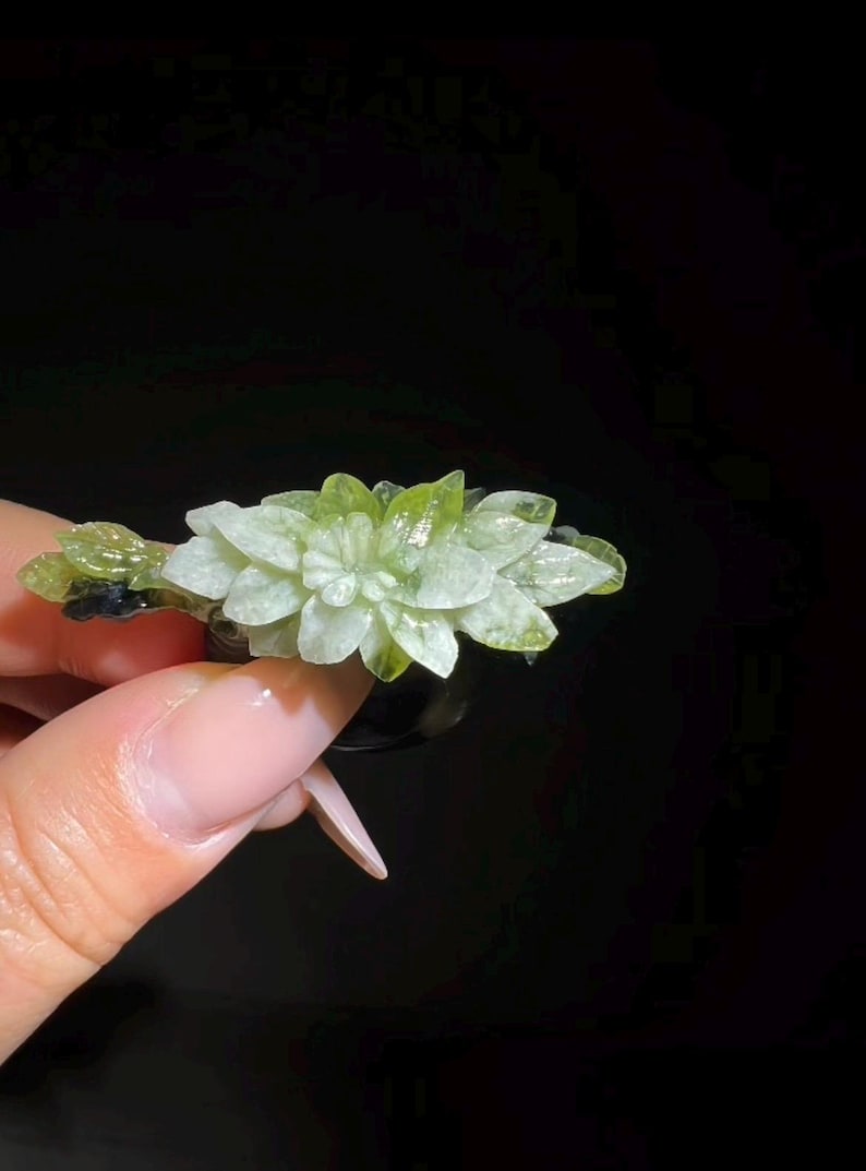 May include: A light green and white floral-shaped crystal. The crystal is held against a black background. The crystal has a detailed, layered design, with a mix of light green and white hues. The overall shape resembles a flower.