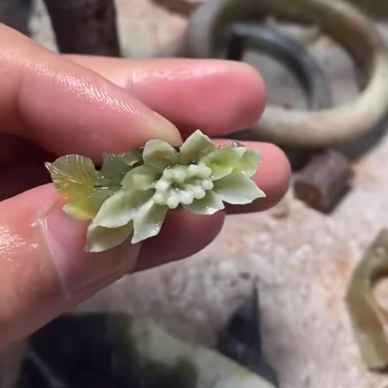 以下が含まれることがあります： 手で持たれた、複数の花びらと葉を持つ花の精巧な彫刻。花は淡い緑色で白いアクセントがあり、葉は黄色がかった色合いです。彫刻はヒスイまたは同様の石で作られているようです。