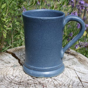 May include: A blue ceramic mug with a handle, on a weathered wooden surface. The mug is cylindrical with a textured surface. The background features green foliage and purple flowers. The mug is approximately 12 cm tall.