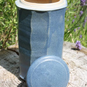 May include: A blue ceramic jar with a lid, set on a wooden surface. The jar has a rectangular shape with a textured surface and a light-colored rim. The lid is round and rests beside the jar. The background features green foliage and purple flowers.
