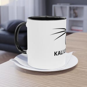 Puede incluir: Taza de cerámica blanca con asa e interior negros, con un diseño gráfico negro y la palabra "KALI" en negro. La taza está sobre un platillo blanco, colocado sobre una superficie de madera.