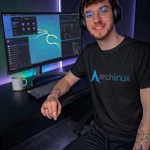 May include: A person wearing a black Arch Linux t-shirt, glasses, and headphones, sitting at a desk with a computer monitor displaying code and the Kali Linux logo. A white mug sits on the desk.