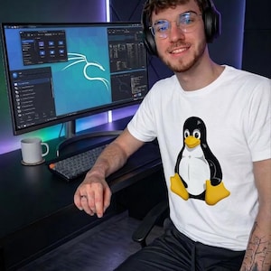Puede incluir: Una camiseta blanca con un pingüino de dibujos animados en negro, blanco y amarillo. La persona lleva auriculares y gafas, y está sentada frente a un monitor de ordenador que muestra interfaces de software.