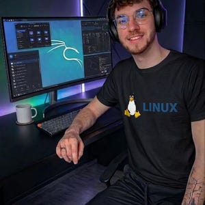 May include: A person wearing a black t-shirt with a penguin graphic and the word "LINUX" in blue. The person is wearing headphones and glasses, and is sitting in front of a computer monitor displaying code and a Kali Linux logo.