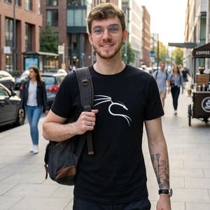 Puede incluir: Una persona con una camiseta negra con un diseño gráfico blanco caminando por una acera de la ciudad. Lleva gafas y una bandolera gris oscuro. Se ven edificios y otras personas al fondo.