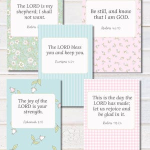 May include: Five decorative cards with inspirational quotes. The cards feature floral, striped, and gingham patterns in pastel colours. Each card has a white frame with a Bible verse and reference. The cards are arranged on a white wooden surface.