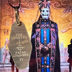 May include: A brass key chain shaped like a hotel room key with the text "The Ministry Papal Chamber IV" on it. The key chain is hanging from a black cord and is in front of a man wearing a black and blue robe with a white mask.