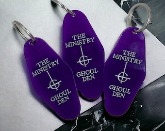 May include: Three purple keychains with silver key rings. Each keychain features a black and white graphic of an upside-down cross with a circle around it and the text "The Ministry Ghoul Den".