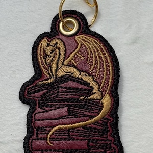 May include: A burgundy and gold dragon keychain. The dragon is embroidered in gold thread, perched atop a burgundy brick structure. The keychain has a black border and a gold-colored keyring.