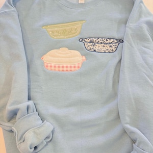 May include: Light blue sweatshirt with three embroidered casserole dish designs. The dishes are in shades of green, white, pink, and blue. The sweatshirt has long sleeves and a crew neck.