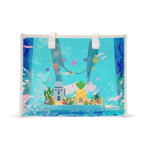 Può includere: Una borsa tote trasparente con una vivace scena sottomarina. Presenta una casa ad ananas, una testa di pietra e coralli colorati. La borsa ha manici bianchi e un bordo bianco, su uno sfondo blu e turchese.