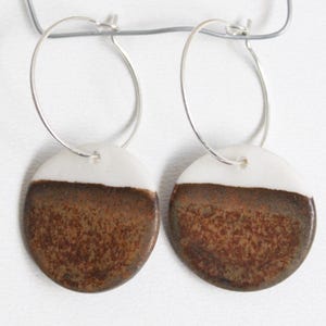 May include: A pair of hoop earrings featuring circular ceramic pendants. Each pendant has a smooth white upper section and a textured, mottled brown lower section. The pendants are suspended from thin, silver-toned metal wire hoops.
