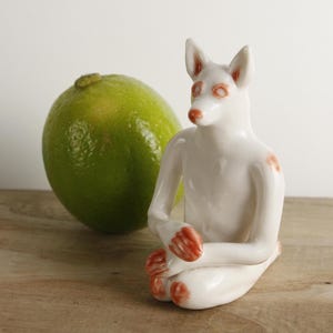 May include: A small white ceramic figurine of a seated anthropomorphic fox with pointed ears and reddish-orange accents on the eyes, ears, hands, and knees. The fox is positioned next to a single whole green lime for scale and visual contrast.