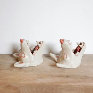 Op de afbeelding: Twee gebroken witte keramische sculpturen in de vorm van open dierenkaken met zichtbare tanden en roze accenten op de neus, ogen en in de mond. De glanzend geglazuurde stukken staan naast elkaar en tonen een gedetailleerde, holle vorm.