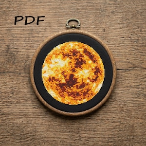 May include: A cross-stitch pattern PDF featuring a circular design of a glowing sun or planet in shades of yellow, orange, and dark brown against a solid black background, displayed within a round wooden embroidery hoop with a metal hanger.