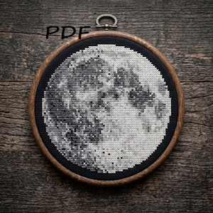 May include: A circular cross-stitch embroidery featuring a detailed greyscale moon set against a solid black background, encased in a round wooden hoop with a metal hanging clasp at the top. The text PDF is visible in the upper left corner.