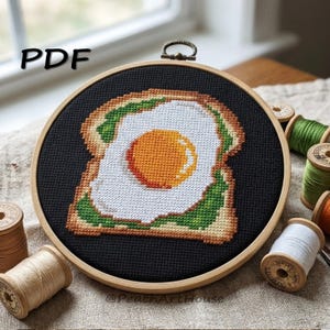 May include: A cross-stitch embroidery hoop featuring a breakfast theme. The design depicts a piece of toast with a fried egg on top, stitched in shades of brown, white, green, and orange. The text "PDF" is in the upper left corner.