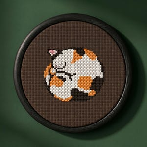 Op de afbeelding: Een cirkelvormig kruissteek borduurwerk met een gestileerde, opgerolde lapjeskat met witte, oranje en zwarte vlekken. Het ontwerp is geborduurd op een donkerbruine stoffen achtergrond en zit in een ronde, zwarte houten lijst.