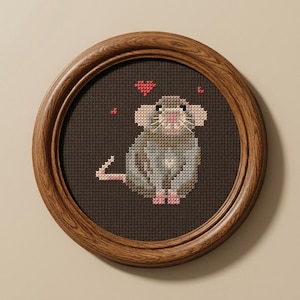 May include: A circular wooden frame containing a cross-stitch embroidery of a grey mouse on a dark brown fabric background. The mouse has pink ears, feet, and tail, with small red heart-shaped stitches floating above its head in the composition.