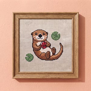 Könnte beinhalten: Ein gerahmtes Kreuzstich-Stickbild mit einem braunen Otter mit weißer Brust, der einen roten Gegenstand hält, umgeben von zwei grünen Seerosenblättern auf hellbeigem Stoff, eingefasst in einen quadratischen Holzrahmen mit natürlicher Maserung.