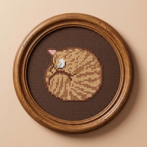 May include: A circular wooden frame containing a cross-stitch embroidery of a curled-up ginger tabby cat. The cat is depicted in shades of tan and brown, resting on a dark brown fabric background. The embroidery shows the cat in a sleeping position.