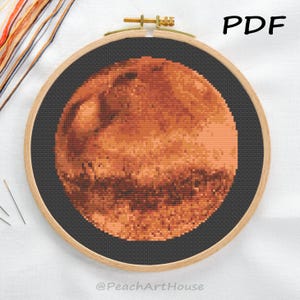 May include: A cross-stitch embroidery of Mars, featuring shades of orange, brown, and tan, set against a black background. The design is framed in a wooden hoop with a brass clasp. The word "PDF" is visible in the upper right corner, and the text "@PeachArtHouse" is at the bottom.