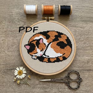 May include: A round wooden embroidery hoop containing a cross-stitch pattern of a sleeping calico cat with black, orange, and white fur. The text PDF is visible. Three spools of thread in white, orange, and black, a pair of metal scissors, and a daisy.