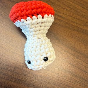 May include: A small, handmade crochet mushroom figure featuring a bright red cap and a cream-colored stem. The stem includes two small, round, black plastic bead eyes positioned near the base, giving the soft, textured fiber object a face.
