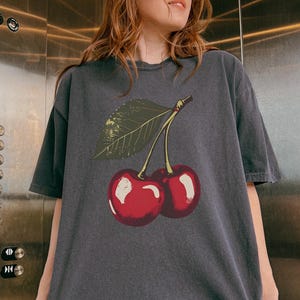 May include: Dark gray t-shirt featuring a vintage-style illustration of two ripe red cherries with a green leaf. The graphic is centered on the front of the shirt, creating a casual and fruity aesthetic.