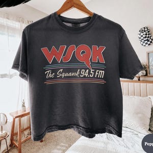 May include: Dark gray t-shirt with the text "WSQK The Squawk 94.5 FM" in red and white lettering. The shirt is hanging on a wooden hanger. The text is layered over a series of horizontal lines in various colors.