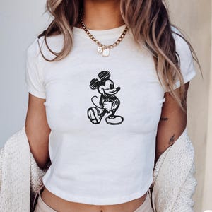 May include: White crop top featuring a black sketch of Mickey Mouse. The classic cartoon character is depicted in a walking pose. The shirt is a short-sleeved style.
