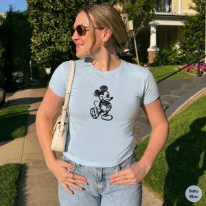 May include: Light blue t-shirt with a black Mickey Mouse graphic. The shirt has short sleeves and a crew neck. The model is wearing blue jeans and sunglasses. The shirt is labeled "Baby Blue".