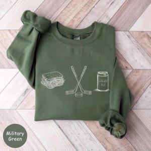May include: Military green sweatshirt with white line art of a sandwich, crossed hockey sticks, a hockey puck, and a can of ginger ale. The text "Ginger Ale" is on the can. The sweatshirt is a solid color with a crew neck and long sleeves.