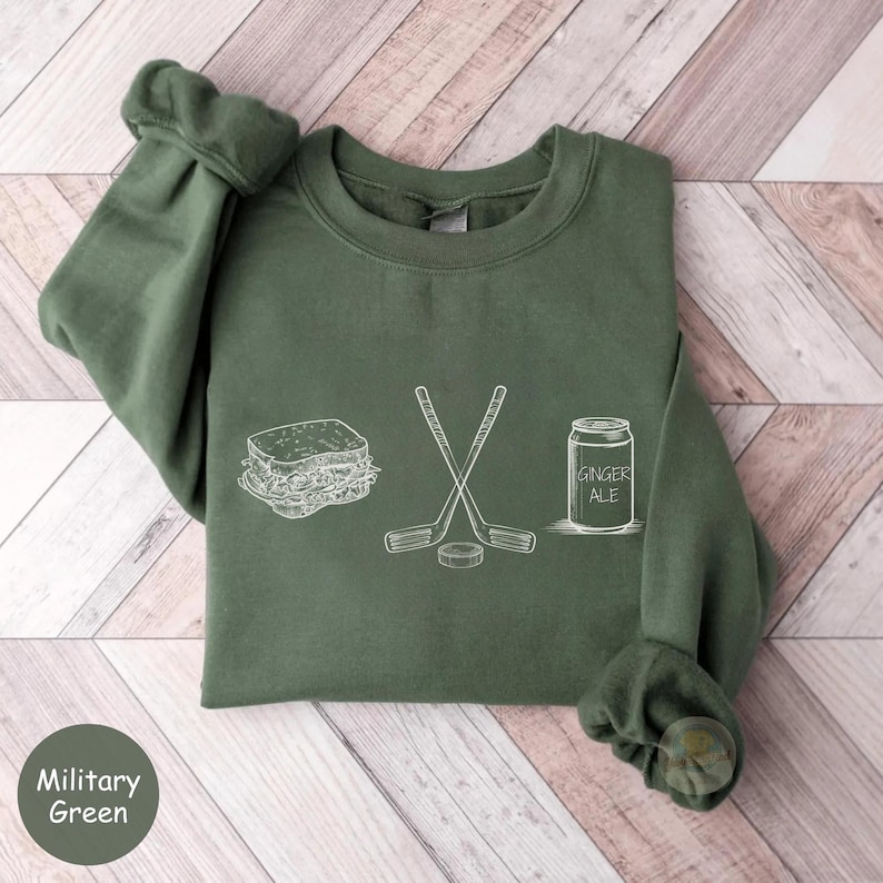 May include: Military green sweatshirt with white line art of a sandwich, crossed hockey sticks, a hockey puck, and a can of ginger ale. The text "Ginger Ale" is on the can. The sweatshirt is a solid color with a crew neck and long sleeves.