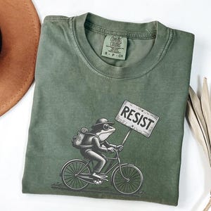 Könnte beinhalten: Olivgrünes T-Shirt mit einer Illustration einer Froschfigur auf einem Fahrrad, die ein "RESIST"-Schild hält. Der Frosch trägt einen Hut und einen Rucksack. Das T-Shirt ist aus weichem Material.