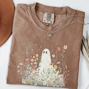 Puede incluir: Camiseta marrón Comfort Colors con un diseño de fantasma rodeado de flores y follaje. Un collar plateado con un colgante descansa sobre la camiseta. La camiseta tiene cuello redondo y mangas cortas.