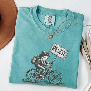 May include: Teal Comfort Colors t-shirt with a frog on a bicycle holding a "RESIST" sign. The frog wears a hat and backpack. A gold necklace is draped across the shirt. The shirt is a Comfort Colors brand.