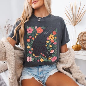 May include: Dark gray t-shirt featuring a floral design with a hand-holding graphic. The design includes colorful flowers and the names "Theresa" and "Emily". The shirt is worn by a person with blonde hair and blue denim shorts.