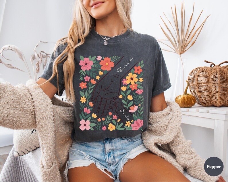 May include: Dark gray t-shirt featuring a floral design with a hand-holding graphic. The design includes colorful flowers and the names "Theresa" and "Emily". The shirt is worn by a person with blonde hair and blue denim shorts.