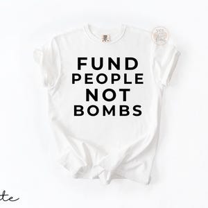 May include: A white short-sleeved t-shirt featuring the black text FUND PEOPLE NOT BOMBS printed in a bold, sans-serif font across the center of the chest. The garment has a crew neckline and a standard fit with a small circular logo in the corner.