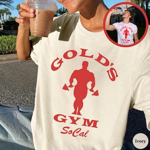 May include: Ivory-colored t-shirt featuring a red graphic of a weightlifter and the words "Gold's Gym SoCal". The shirt has a classic crew neck and short sleeves. The image also shows a person holding a clear plastic cup with a brown beverage.